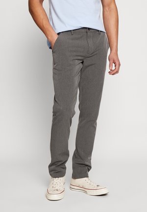 Mand iført grå slim-fit bukser, hvide lærreds sneakers og en lyseblå skjorte, stående med den ene hånd i lommen mod en ensartet baggrund.