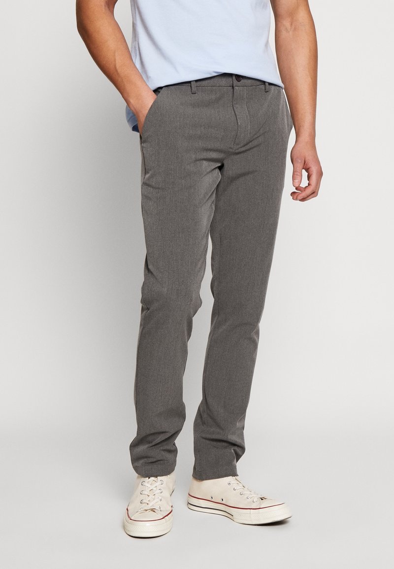 Homme portant un pantalon gris slim fit, des baskets blanches en toile et une chemise bleu clair, debout avec une main dans la poche devant un fond uni.
