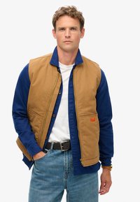 Gilet en toile marron porté sur une chemise bleu à manches longues, avec une fermeture éclair sur le devant, des poignets boutonnés et des poches avant. Un jean en denim décontracté complète le look.