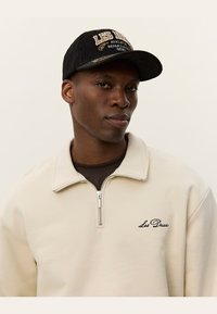 Sudadera con cremallera color beige claro, con un logo bordado, llevada sobre una camiseta oscura, combinada con una gorra de pana negra que presenta texto y detalles.