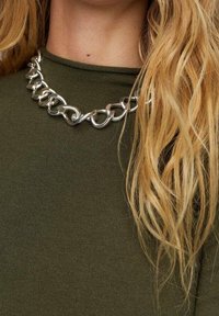 Collier à grosse chaîne en argent porté sur un haut col montant vert olive, avec de longs cheveux blonds ondulés couvrant partiellement le côté droit.