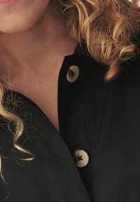Zwarte stoffen shirt met knoopsluiting, met een grote ronde knoop met een kruispatroon en een kraag. Krullend haar is gedeeltelijk zichtbaar.