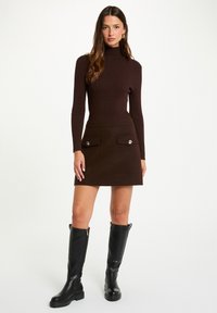 Maglione collo alto a coste marrone abbinato a una gonna marrone con due tasche frontali e dettagli in bottone argentato, completato da stivali neri al ginocchio.