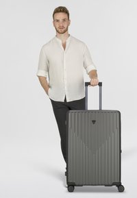 Valise rigide grise avec texture à stries verticales, poignée rétractable et quatre roulettes. Logo triangulaire distinctif à l'avant.