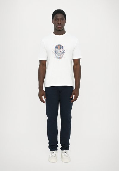 Camiseta blanca de manga corta con un colorido diseño de calavera en el frente, combinada con pantalones azul marino oscuros y zapatillas blancas.