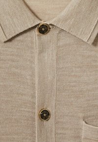Camisa polo beige claro con un collar clásico, tejido texturizado y un botón oscuro prominente con un borde dorado, realzado por un detalle de bolsillo.