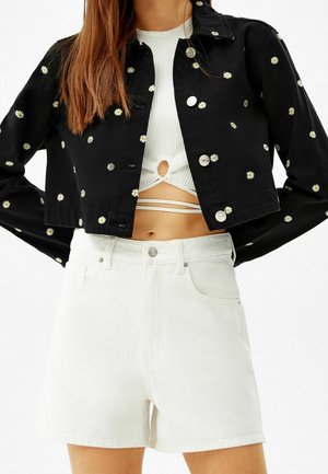 Vrouw draagt een zwart cropped jack met madeliefjesprint over een wit cropped topje met uitsnijding en witte high-waisted denim shorts.