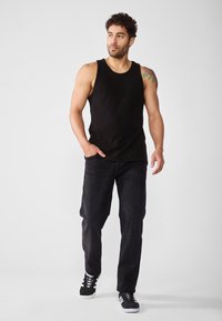 Canottiera nera a coste abbinata a jeans scuri e sneakers nere. Il modello ha i capelli ricci e visibile un tatuaggio sul braccio.