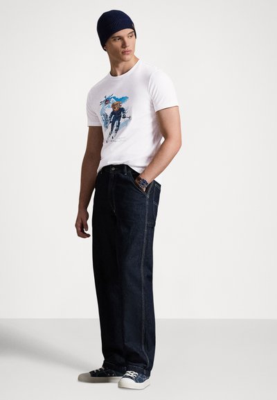 Bijela majica s obojenim printom, tamne traperice i tamnoplava kapa. Model nosi plave tenisice s bijelim đonom.