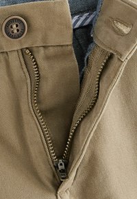 Fermeture éclair en tissu beige avec un bouton en bronze, présentant une fermeture éclair en métal doré. La doublure intérieure possède un motif textile rayé.
