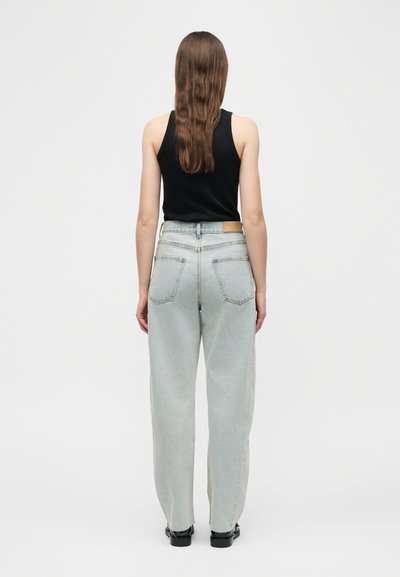 Γυναίκα με μακριά καστανά μαλλιά, φορώντας μαύρο τοπ χωρίς μανίκια, ανοιχτόχρωμο τζιν ψηλόμεσο παντελόνι και μαύρα παπούτσια, όρθια με την πλάτη προς το θεατή.