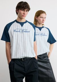 Νεαρός άνδρας και γυναίκα φορούντα λευκά και μπλε μπλουζάκια με το ίδιο λογότυπο, στέκονται μπροστά σε απλό ανοιχτό φόντο.