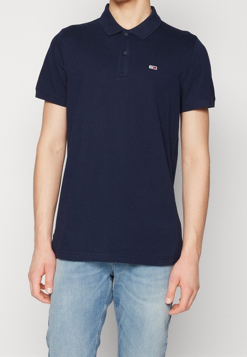Homme portant un polo à manches courtes bleu marine avec un petit logo sur le côté gauche de la poitrine et un jean bleu clair.