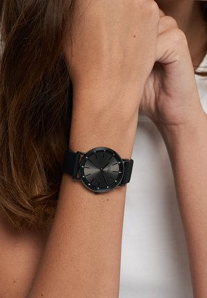 Schwarze Analoguhr mit rundem Zifferblatt, matter Oberfläche und minimalistischem Design. Verfügt über ein Silikonarmband und Kristallakzente um das Zifferblatt.