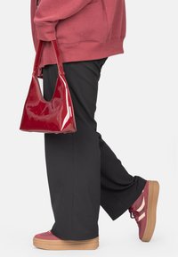 Rote Lackleder-Handtasche mit dreieckiger Form und breitem Ausschnittgriff, kombiniert mit schwarzen Hosen und roten Sneakers mit Plattformsohle.
