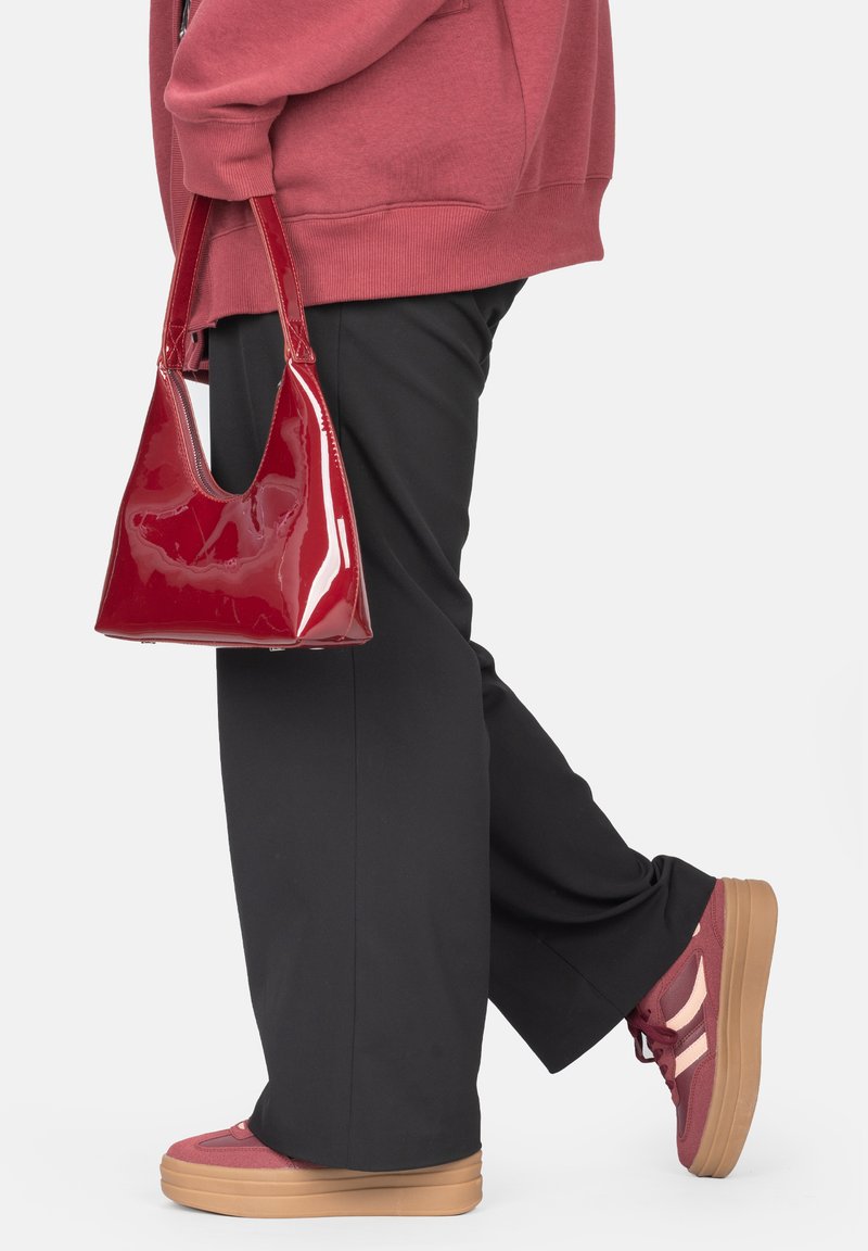 Rote Lackleder-Handtasche mit dreieckiger Form und breitem Ausschnittgriff, kombiniert mit schwarzen Hosen und roten Sneakers mit Plattformsohle.