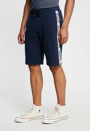 Mężczyzna ubrany w granatowe szorty z białym paskiem "Hollister" po bokach oraz białe wysokie trampki, stojący na jednolitym tle.