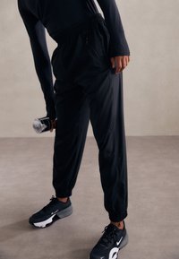 Black athletic trousers with elastic waistband, zippered pockets, and tapered ankles. Worn with black sneakers and a dumbbell in hand.