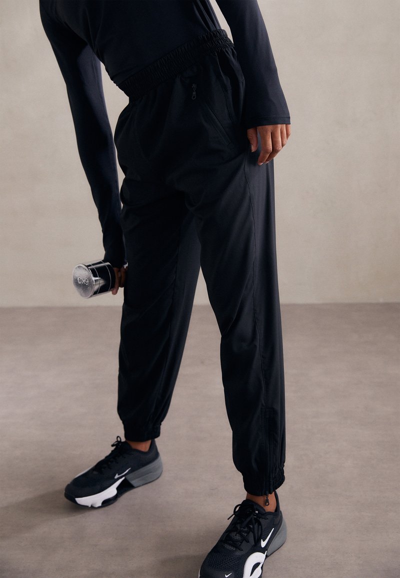 Black athletic trousers with elastic waistband, zippered pockets, and tapered ankles. Worn with black sneakers and a dumbbell in hand.