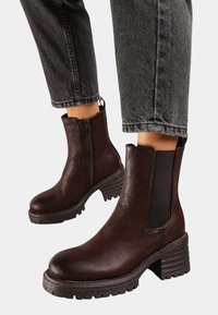 Bottines en cuir marron avec une finition texturée, des panneaux latéraux élastiques et des talons en caoutchouc épais. Portées avec un jean noir à revers.