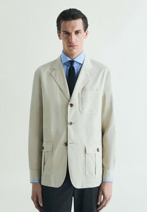 Hombre con blazer beige de tres botones, camisa azul y corbata negra, de pie frente a un fondo claro y liso.