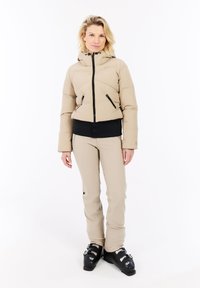 Kvinde iført en beige polstret jakke og bukser med sorte skistøvler, stående mod en hvid baggrund.