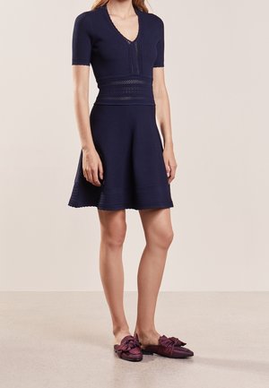 Granatowa dziergana sukienka z krótkimi rękawami, dekoltem w serek i teksturowanym wzorem; dopasowana góra rozwija się w rozkloszowaną spódnicę. Połączona z ciemnofioletowymi butami akcentowanymi kokardą.