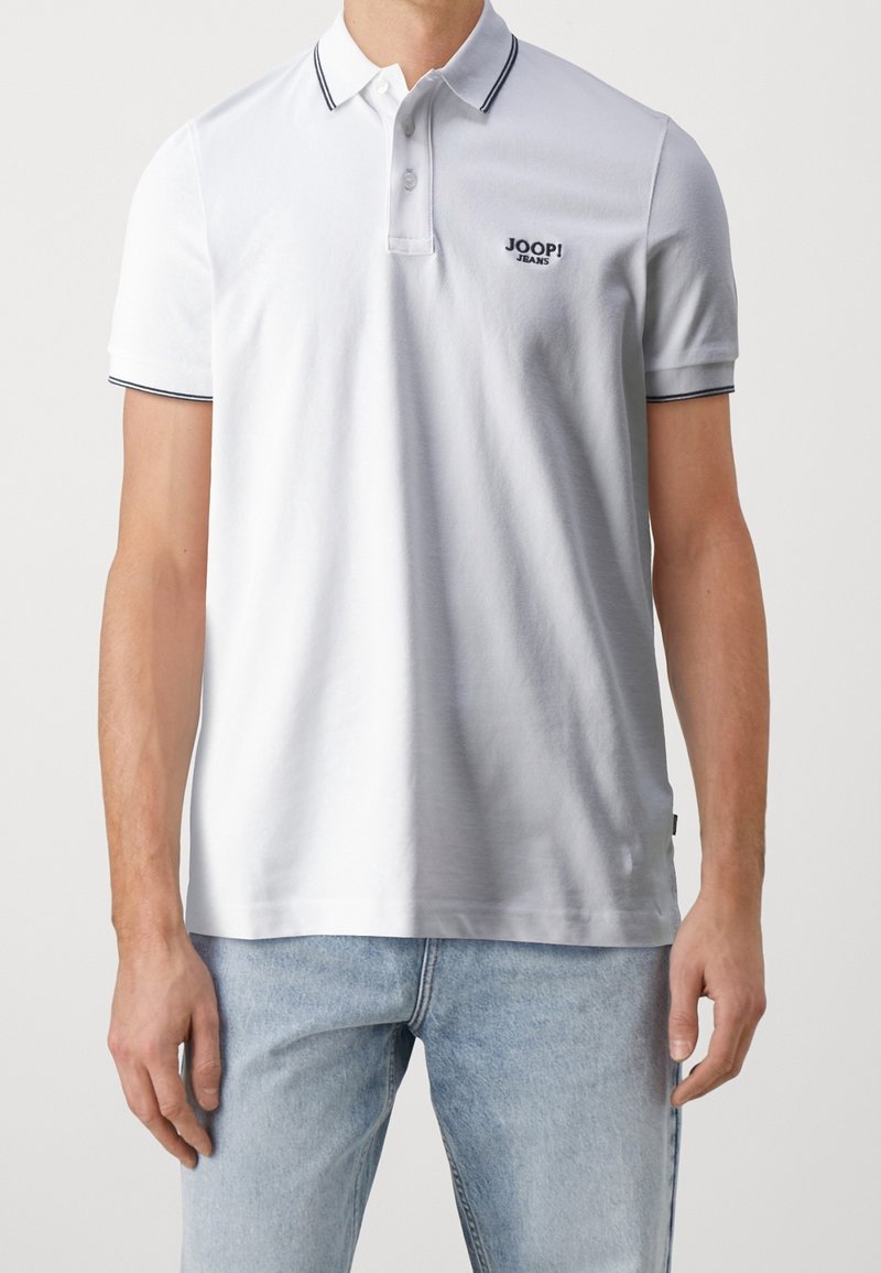 Homme portant un polo blanc à manches courtes avec bordure noire et logo "JOOP! JEANS", associé à un jean bleu clair.