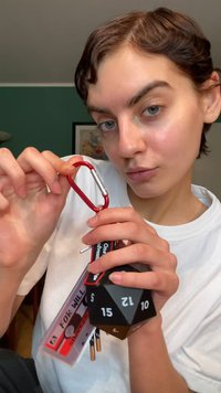 Rode aluminium karabijnhaak in de hand, bevestigd aan een zwart veelvlakdobbelsteen met witte cijfers, samen met sleutelhangers in rood en zwart.