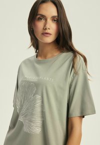 Jasnozielona bawełniana koszulka z dużą białą grafiką rośliny oraz napisem "Świat Roślin" w swobodnym fasonie z krótkimi rękawami.