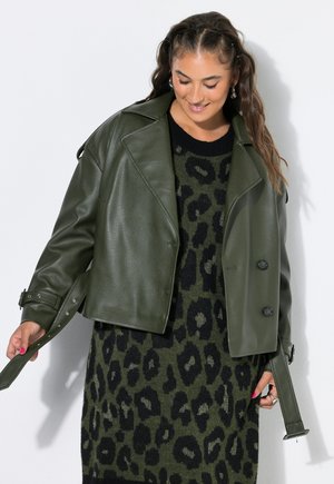 Kvinde med langt bølget hår iført en mørkegrøn læderjakke over en mørkegrøn og sort kjole med leopardprint, der holder jakkeens bæltestraps.