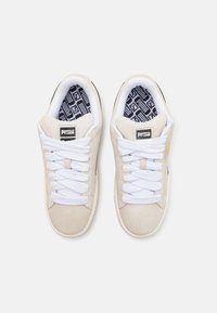 Kremowe zamszowe sneakersy z białymi sznurówkami, z czarną podeszwą i wkładką w geometryczny wzór. Logo na języku i akcenty po bokach.