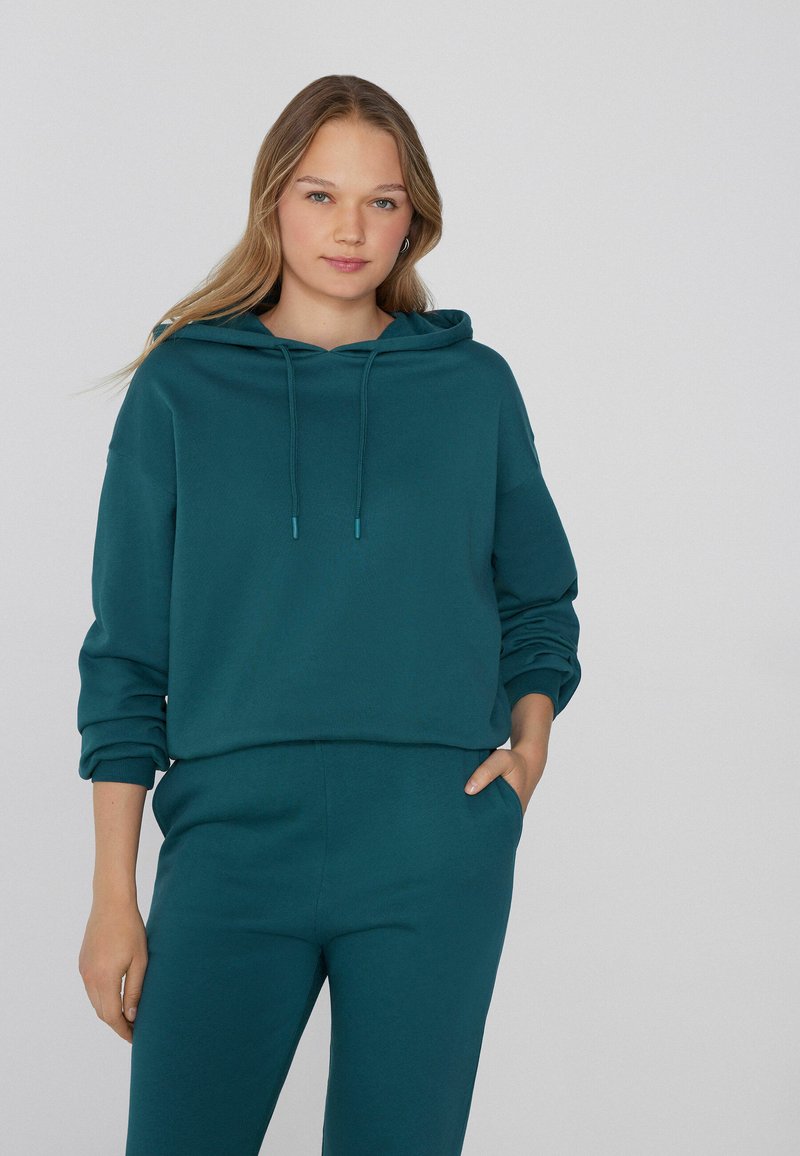 Sudadera azul verdosa con capucha ajustable, puños acanalados y un corte relajado, combinada con pantalones a juego. Textura suave y lisa, diseño casual.