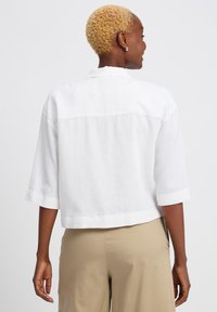 Witte, verkorte linnen shirt met korte mouwen, voorzien van een achterste schoudernaad en contrasterende textuurstof. Draagt samen met lichtbeige broek.