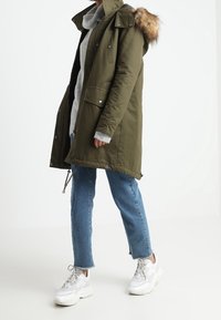Parka vert olive avec capuche doublée de fourrure, équipée d'une fermeture éclair et de boutons-pression, deux poches avant, portée avec un pull gris et un jean bleu.