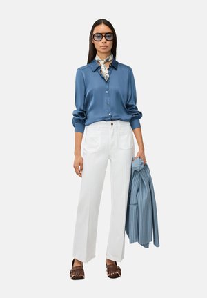 Donna che indossa una camicetta di raso blu, pantaloni bianchi a gamba larga, mocassini marroni, occhiali e sciarpa al collo, che tiene in mano un blazer blu a righe sottili.