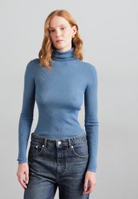 Blue ribbed turtleneck sweater with long sleeves, fitted design; paired with high-waisted dark denim jeans. Plain background.