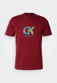 T-shirt à manches courtes couleur marron avec col rond, présentant un logo "CK" bleu et vert et un texte "Calvin Klein" au centre devant.
