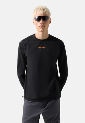 Lyshåret person iført sort langærmet skjorte med "FIRE+ICE"-logo, mørke bukser og store sorte solbriller, stående mod en grå baggrund.