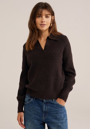Frau mit braunen Haaren, die goldene Creolen, einen dunkelbraunen Strickpullover mit Kragen und blaue Jeans trägt, mit einer Hand in der Tasche.