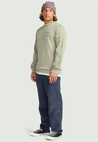 Homem a usar um sweatshirt verde claro, uma touca roxa, calças azuis escuras e sapatilhas pretas, de pé contra um fundo claro e liso.