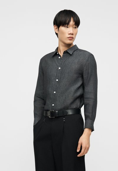 Νεαρός άντρας που φοράει σκουρόγρινο πουκάμισο με κουμπιά, μπαίνει μέσα σε μαύρο παντελόνι με μαύρη ζώνη, στέκεται με τα χέρια στις τσέπες μπροστά σε απλό φόντο.