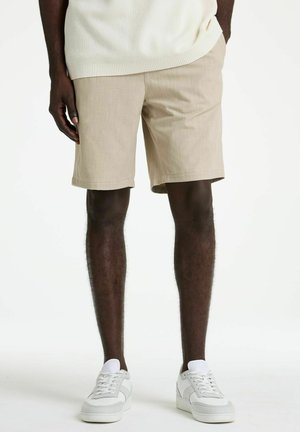 Mann trägt beige Shorts, weiße Sneakers und einen cremefarbenen Strickpullover, steht mit einer Hand in der Tasche vor einem weißen Hintergrund.