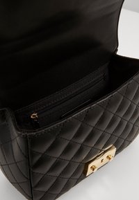 Borsa a tracolla nera trapuntata con hardware dorato. L'interno presenta una tasca con zip e una fodera in tessuto morbido. Forma squadrata con design strutturato.