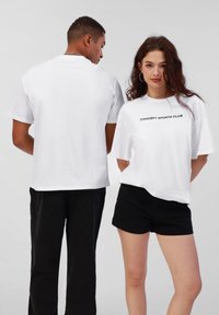 Hvite bomulls t-skjorter med svart "CONCEPT SPORTS CLUB" trykk på fronten, parret med svarte shorts og bukser, vist fra front og bak.