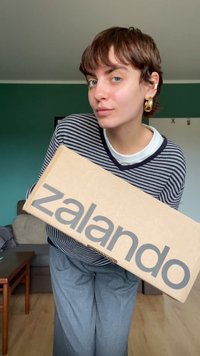 Caja de cartón con "zalando" impreso en gris en el lado, sostenida por una persona que lleva una camiseta de rayas y pantalones grises holgados, en interiores.