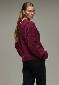 Bordeauxroter, gestrickter Pullover mit Zopfmuster, schwarzen Bandakzenten an den Ärmeln und lockerem Schnitt, kombiniert mit dunklen Jeans.