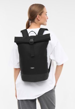 Woman with light brown hair in a ponytail wears a white shirt and gray pants, carrying a large black backpack with a roll-top and buckle.