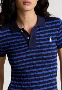 Mujer vistiendo una camiseta de polo de punto a rayas en azul marino y azul brillante, con cuello y un logotipo bordado en blanco en el pecho.