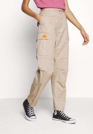 Pantalons cargo beige avec plusieurs poches, taille haute et un petit patch logo orange. Tissu texturé et coupe droite, associés à des baskets noires.